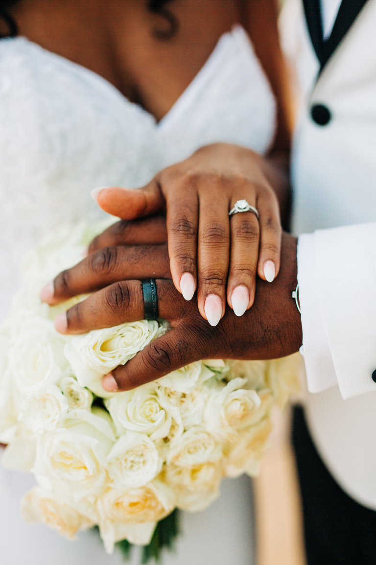 Black Bride and Groom Hands with Wedding Bands Over White Roses Bouquet